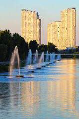Fototapeta premium Scenic sunset landscape of Rusanivka neighborhood at sunny day. Fountains in the city channel. Modern skyscrapers in the background. Landscape during the autumn season. Kyiv, Ukraine