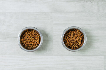 Two metal bowls with dry cat food on white wooden floor.