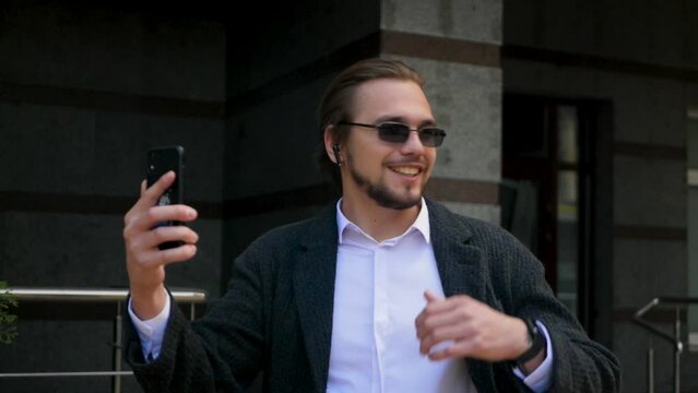 Wealthy man with long hair having video call on his phone outside. Successful businessman participating in outside conference call with colleagues and business partners. Video call with earphones
