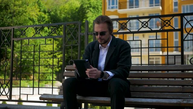 Attractive wealthy man sitting on bench and working on his tablet in slow-motion. Stylish caucasian male in sunglasses trying to solve problem on tablet. Serious modern businessman scrolling on tablet