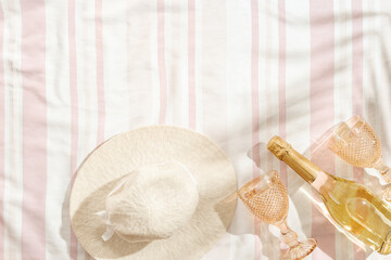 Obraz na płótnie Canvas Summer pastel background with sun hat, color wine glasses, bottle white wine, sparkling drink on striped beach towel as background, shadow of palm leaf. Concept of summer hot day on beach