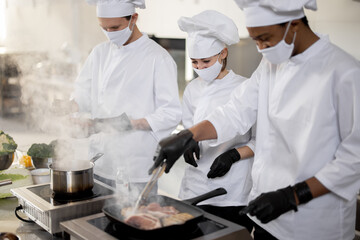 Multiracial team of cooks in uniform and face masks cooking meals for a restaurant in the kitchen. Concept of teamwork at restaurant during pandemic. Latin, Asian and European guys cooking together