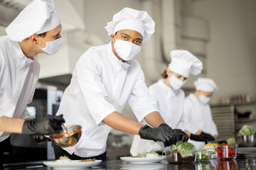 Multiracial group of cooks finishing main courses while working together in the kitchen. Cooks wearing uniform and face mask. Team prepares meals for the restaurant during pandemic