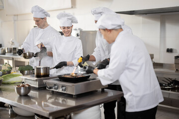 Multiracial team of professional cooks in uniform preparing meals for a restaurant in the kitchen. Latin guy frying meat, european cooks making sauce and asian chef managing the process. Teamwork and