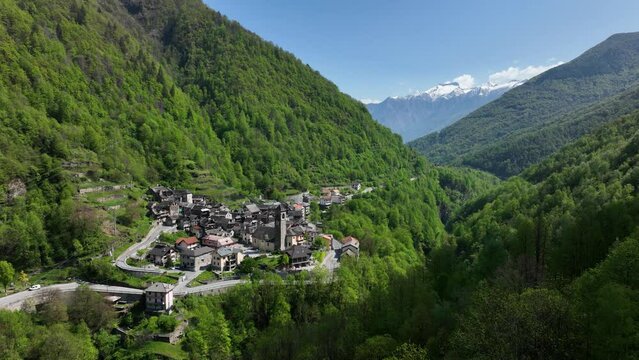 Flight above treetops toward small Italian alp village in lush valley; drone