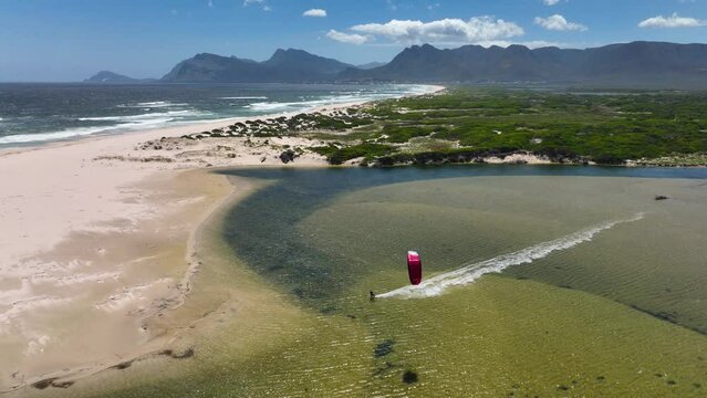 Aerial wide shot of a kite surfer racing and doing stunts in calm water near near the ocean on a sunny day with majestic mountain range in the background near Hermanus South Africa