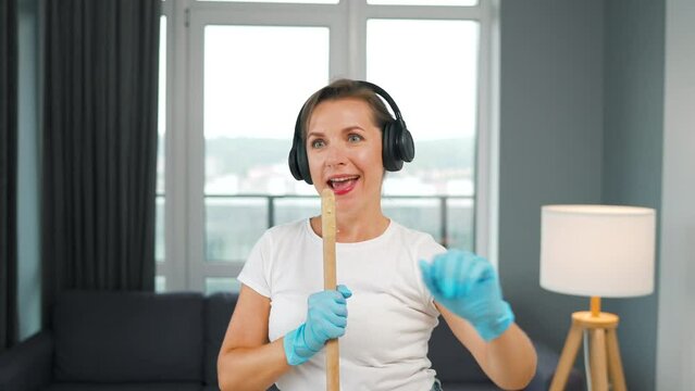 Woman in headphones cleans the house and have fun singing with a broom like a star at a concert.