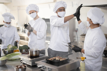 Multiracial team of cooks giving five with hands each other, while cooking in professional kitchen. Concept of teamwork and success at restaurant business. Cooks wearing face masks and protective