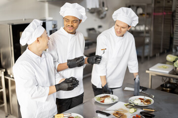 Three cooks with different nationalties tasting cooked meals, inventing new dishes for a menu. Asian chef with Latin and European cooks in uniform working together. Concept of teamwork and high