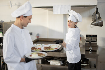 Chef standing with ready meals, talking with female cook in the kitchen. Happy caucasian cook working at restaurant. Concept of professional occupation and teamwork