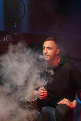 Obraz premium a young man smokes a hookah in a dark hookah, exhales a large cloud of white smoke.