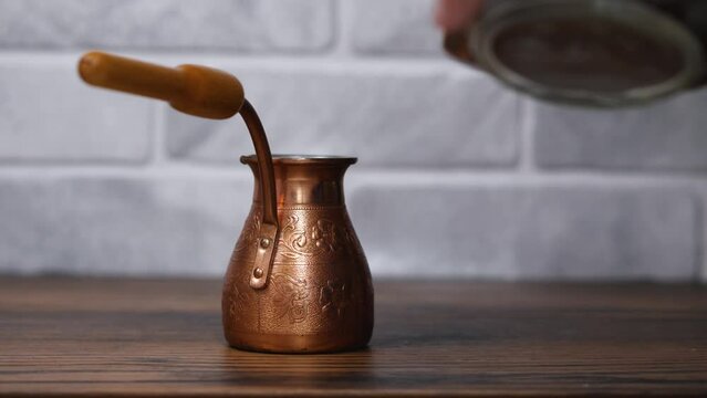 A man's hand with a wooden spoon pours fresh ground coffee into a small copper cezve. In the background is a bright stone wall. The concept of Turkish coffee making