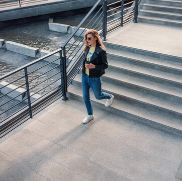 Teen Walk Style. Young Blonde Girl In Jeans And Black Leather Jacket Is Going Down The Gray Stairs With Coffee In Hand And Looks Apart In Sunglasses At Street . Top View. Lifestyle Concept, Free Space