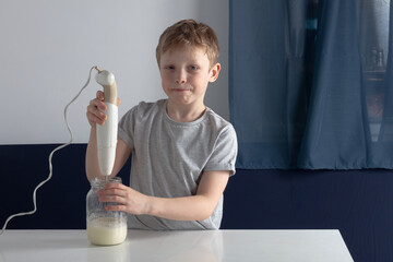A Caucasian boy whips cream from cream or egg white with a blender in a glass jar in the kitchen at...