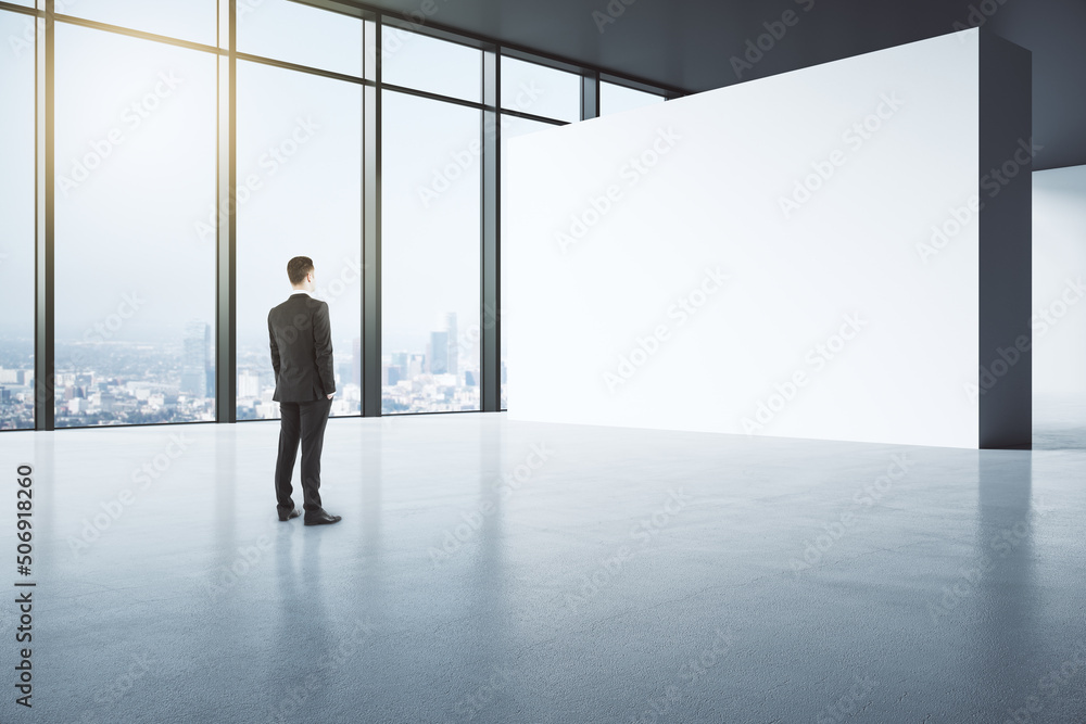 Poster Perspective view businessman in black suit back view looking at blank light partition in empty spacious gallery hall with glossy concrete floor and city view from huge window, mock up