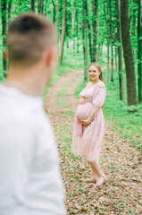 Pregnant woman in a pink dress and her husband look at each other in the green forest