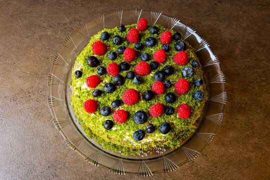 Top view of a round homemade fruit cake with red raspberries, blueberries and green top made of spinach placed on glass dish on brown surface.