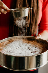 A woman is holding a fresh home made cake and sprinkling powdered sugar on top of it. The movement is blurred. Preparing for a birthday at home. The sponge cake is still in the cake tin. 
