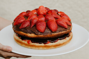A home made sponge cake with fresh strawberry and chocolate cream on a white plate held by a hand. Close up of a delicious summer birthday cake. Two layers, jam and cream in between. Simple decoration