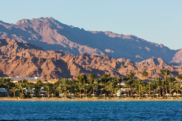 Fototapeta premium The coast of Lagoon beach with hotels, palm trees and mountains. View across the sea. Dahab, Egypt