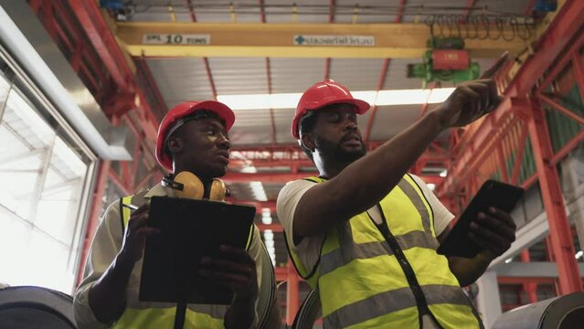 Two African man workers in uniform wearing safety helmet holding tablet and clipboard check inventory stock in factory. Professional industry engineer are inspection and quality control in warehouse. 
