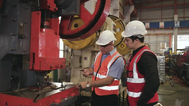 Senior engineer wears safety goggles and helmet standing holding magnet with training young engineer operate hydraulic press machine in factory. Two industry maintenance workers working in warehouse. 