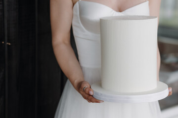 The bride in a white dress holds a white wedding cake. Close-up