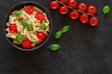 Fusilli pasta cooked with tomato and basil, cherry tomatoes branch on dark background, top view, space to copy text.