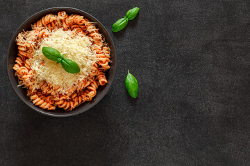 Fusilli pasta cooked with tomatoes sauce, basil and grated parmesan cheese on dark background, top view, space to copy text.