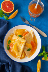Delicious crepes suzette with orange syrup and slices fruits on a plate on a blue concrete background, top view
