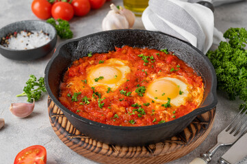 Shakshuka in a frying pan on a gray concrete background with ingredients. Eggs cooked in tomato sauce with spices and fresh parsley on top. Copy space.
