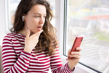 Surprised young woman looking at mobile phone screen with open mouth standing near window indoor, wearing red striped t-shirt. Received unexpected email notice, unbelievable news, shocked content