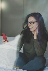 Obraz premium Pretty brunette woman using smartphone sitting on sofa at home. Checking social media, chatting with friend,reading news, watching online webinar,shopping, browsing website,ordering delivery.Vertical