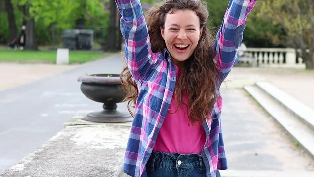 Happy woman celebrating success, victory standing alone in park outdoors. Excited curly student doing winner gesture, jumping, waving her hands, looking at camera. Feeling euphoria. Slow motion