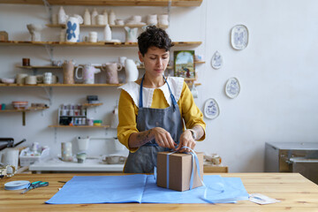 Woman working in craft ceramics shop or studio packing pottery in gift box for courier delivery to...