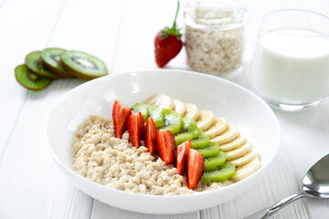 Bowl of oatmeal with fresh fruits, strawberry, banana, kiwi with glass of milk and jar with oats on background. Recipe of dairy breakfast, fitness food.