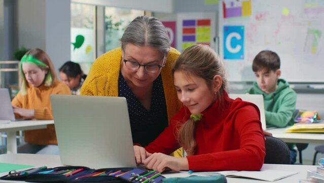 Mature teacher and teenage schoolgirl do exercise on laptop in classroom