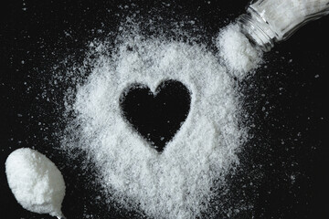 Heart made of salt, which is scattered on the table. Nearby is a metal spoon with salt and an open salt shaker with which salt is poured out - obrazy, fototapety, plakaty