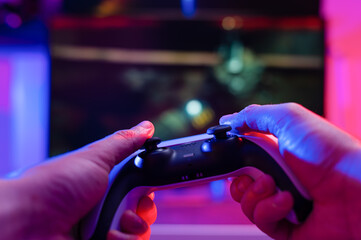Gamepad in the hands of a gamer on a technological background. Plays online video games with friends. Fun pastime, adventure games, youth culture. Close-up.