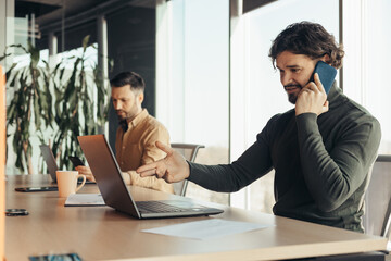 Handsome young businessman using laptop, speaking on smartphone, working with male colleague at modern office