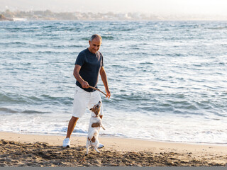 Elderly 60-years old man running with small cute dog jack russell terrier by the sea