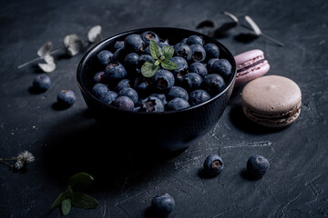 close-up of fresh blueberries in a black bowl with macaroons around on a black slate. the macaroon a small French almond cake. close-up view