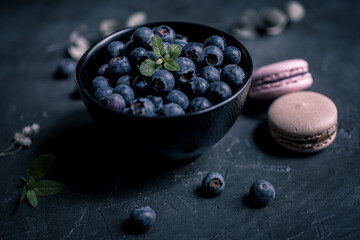close-up of fresh blueberries in a black bowl with macaroons around on a black slate. the macaroon...