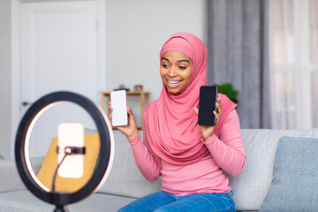 Cheerful muslim black blogger filming video review of two new smartphones, talking to cellphone...