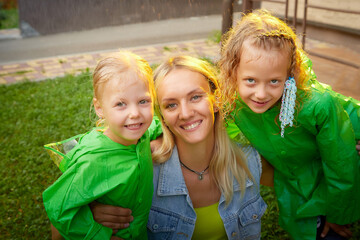 Two cute little blonde girls sisters in green raincoats and mother on a green lawn under rain drops in a summer sunny day near rural house in a village. Family having rest and fun in good spring day