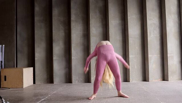 Young girl teenager doing stretching in gym. Blond woman gymnast ballerina dancing at wooden bench. Girl in pink sports suit doing acrobatic figures and string. Long-haired 16-year- old girl do sports