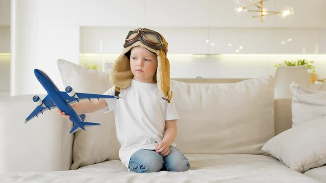 Child dreams of flying an airplane, traveling by air. Cheerful child plays with modern airplane sitting on couch of house. Boy in pilot hat controls toy plane flying in different directions.