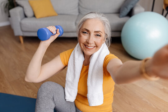 Excited senior woman taking selfie while exercising with dumbbells at home, sitting on yoga mat
