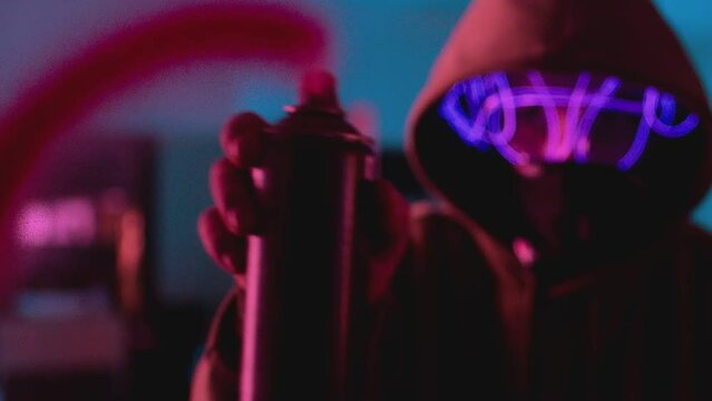 Front view young boy on hoodie and protective mask spraying paint from aerosol on camera . Led neon lights . Close up unrecognizable face with respirator . Art vandalism activist concept Slow motion 