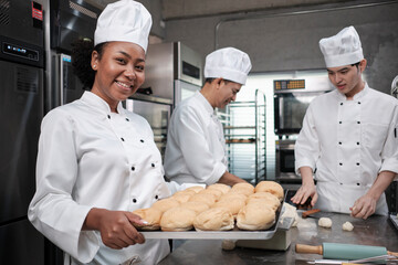 Portrait of African American female chef in white cooking uniform looking at camera with cheerful smile and proud with tray of bread in kitchen, pastry foods professional and fresh bakery occupation.